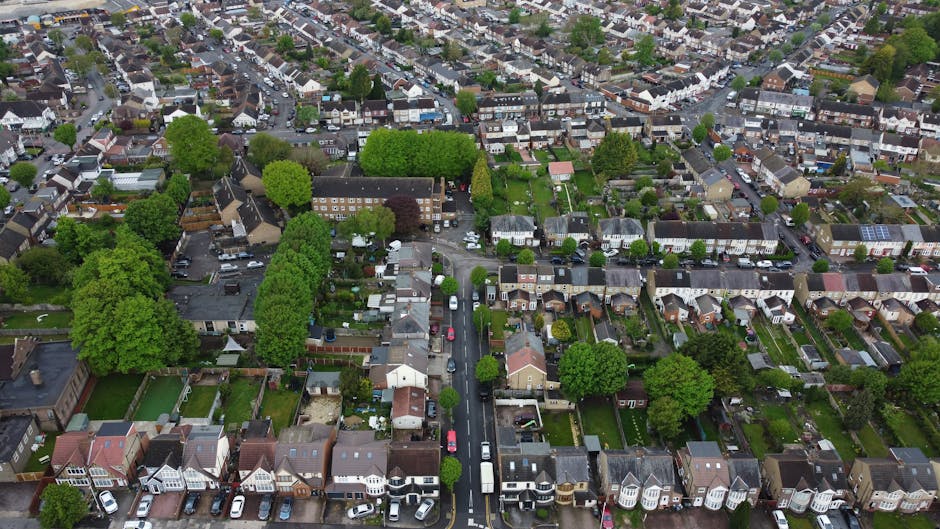 An aerial view of a residential suburban area featuring a mix of houses, gardens, and trees. The gardens show well-maintained lawns, flower beds, and hedges, with some properties bordered by small fen